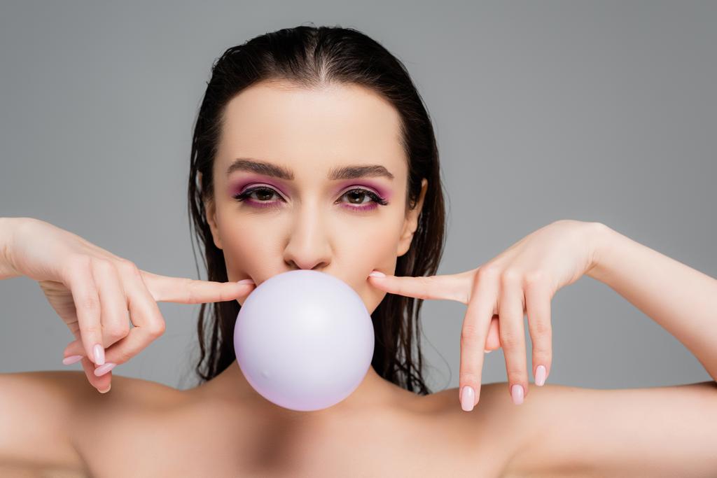 brunette woman with bare shoulders blowing bubblegum and pointing with fingers at cheeks isolated on grey - Photo, Image