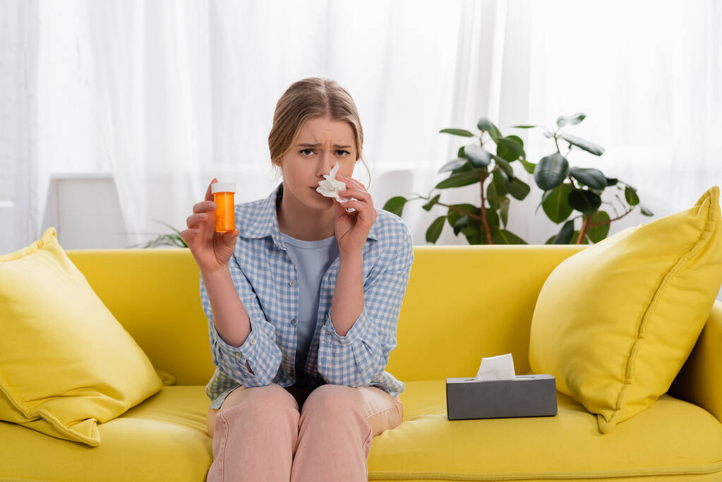 Κουρασμένη γυναίκα με αλλεργία κοιτάζοντας κάμερα, ενώ κρατώντας χάπια και χαρτοπετσέτα στον καναπέ  - Φωτογραφία, εικόνα