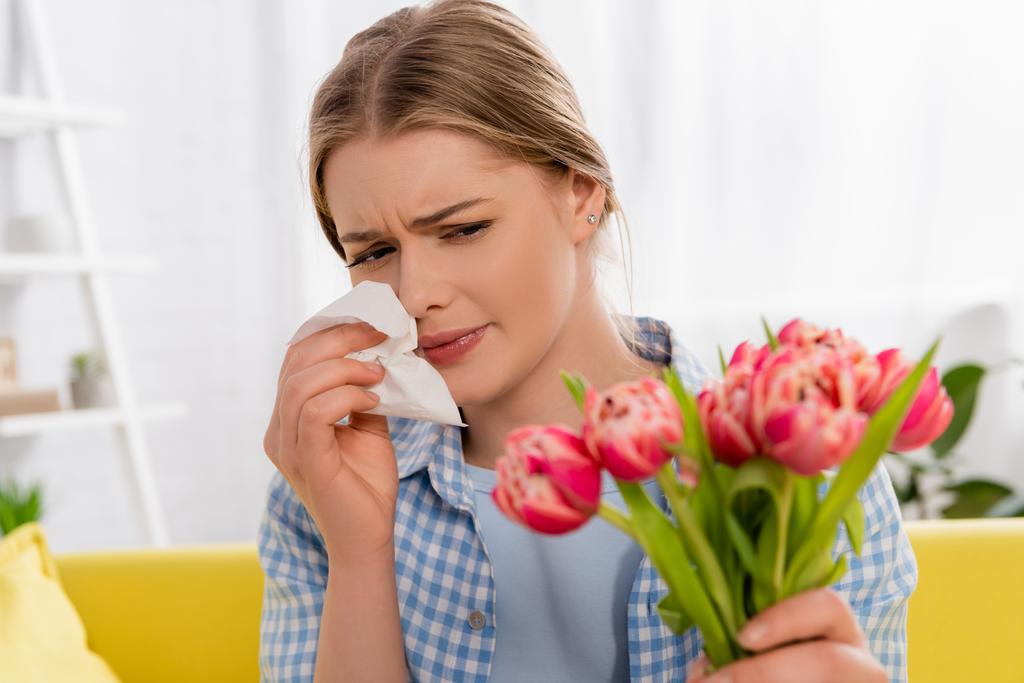 Tired woman holding napkin while suffering from allergy near tulips on blurred foreground  - Photo, Image