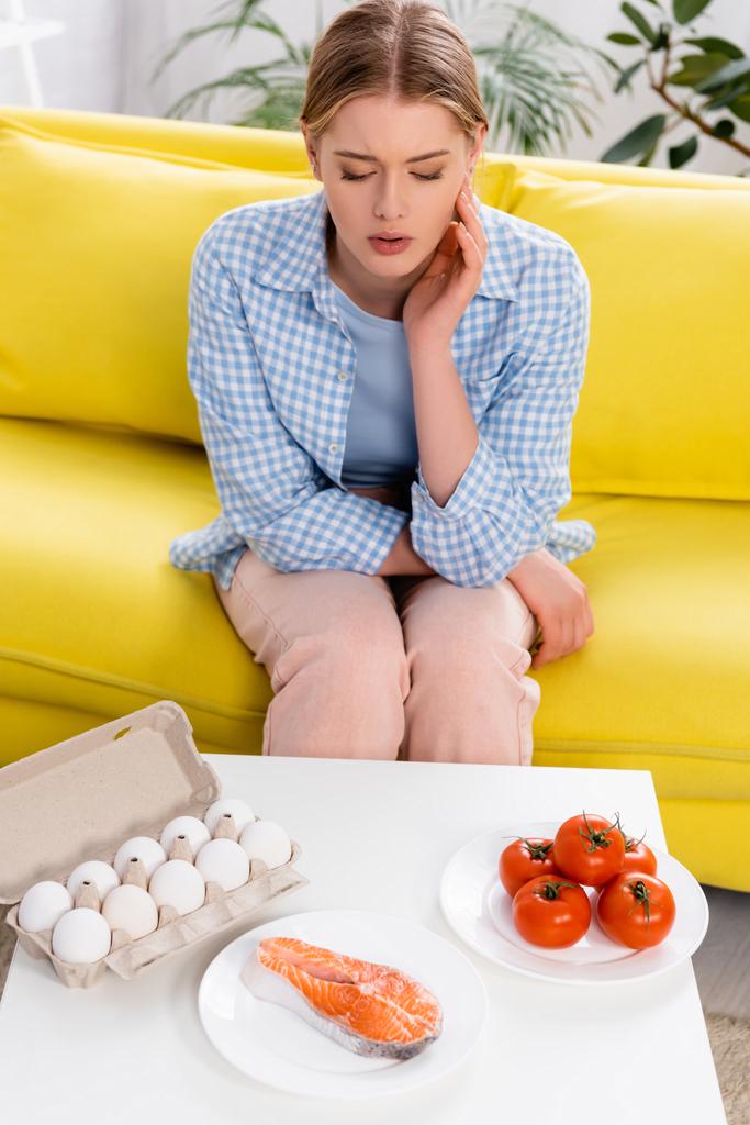 Salmon, eggs and tomatoes near woman with allergy on couch  - Φωτογραφία, εικόνα