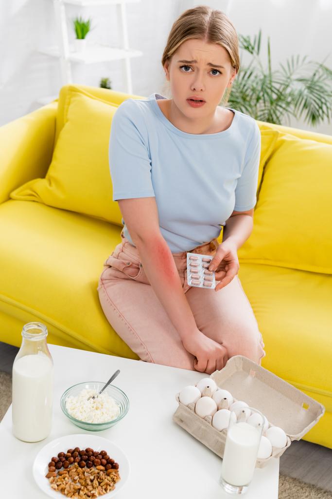 Upset woman with allergy reaction holding pills near food, nuts and milk  - Foto, immagini