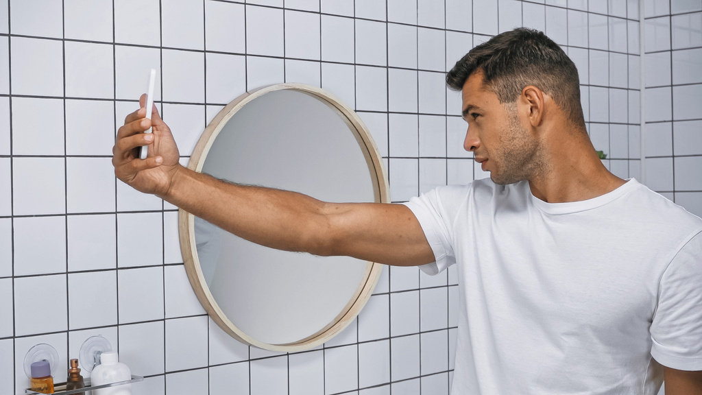 fiatal férfi fehér pólóban csinál selfie mobiltelefon a fürdőszobában - Fotó, kép