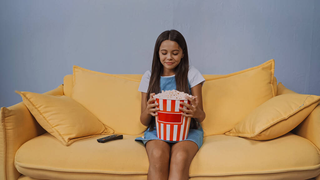 happy kid looking at bucket with popcorn while sitting on sofa  - Photo, Image