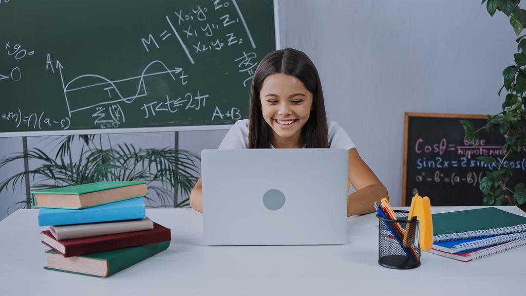 счастливая школьница улыбается во время электронного обучения и смотрит на ноутбук - Фото, изображение