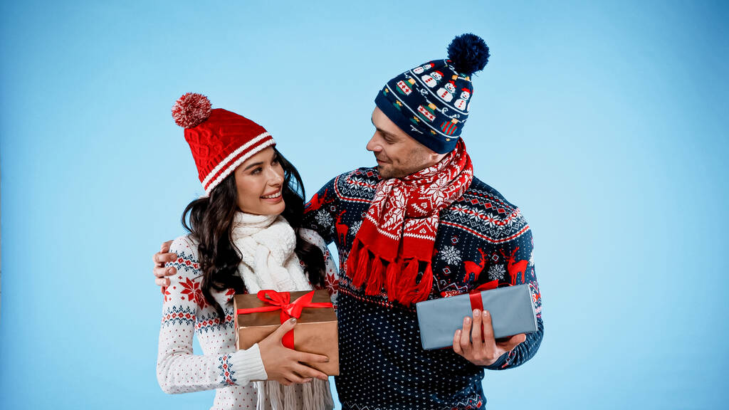 Veselý pár v kloboucích, šátky a svetry drží dárky na modrém pozadí - Fotografie, Obrázek