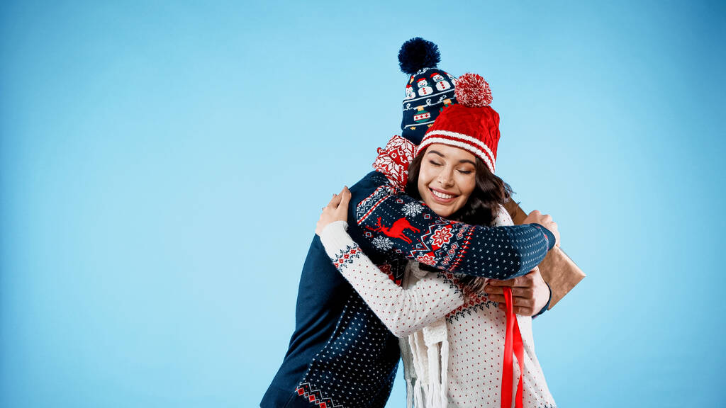 Lächelnde Frau mit Hut und Pullover umarmt Freund mit Geschenkbox auf blauem Hintergrund - Foto, Bild