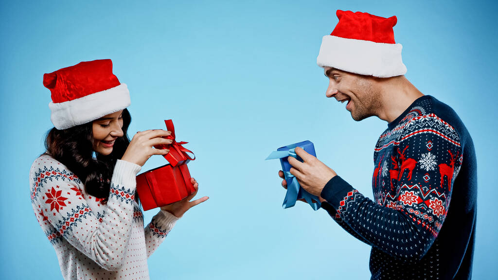 Coppia allegra in cappelli di Babbo Natale e maglioni in possesso di regali su sfondo blu - Foto, immagini