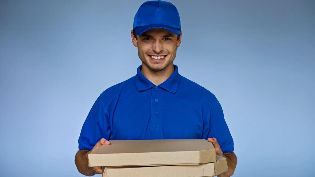 joyful delivery man holding pizza boxes while smiling at camera isolated on blue - Photo, Image