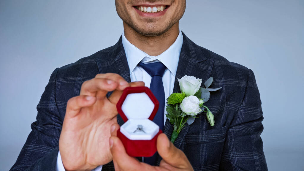 oříznutý pohled na šťastného ženicha zobrazující šperkovnici se snubním prstenem na rozmazaném popředí izolované na modré - Fotografie, Obrázek