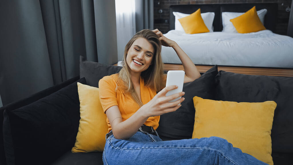 cheerful woman taking selfie on couch in hotel room - Photo, Image
