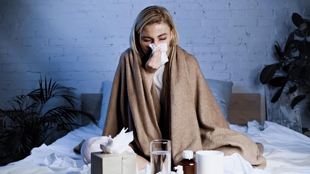 sick woman sneezing into napkin while sitting on bed near medications - Photo, Image