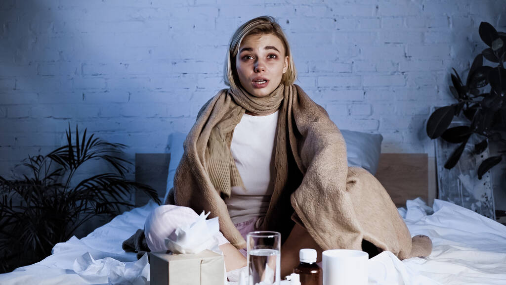 mulher doente sentado na cama sob cobertor quente perto da mesa com bebidas e medicamentos - Foto, Imagem