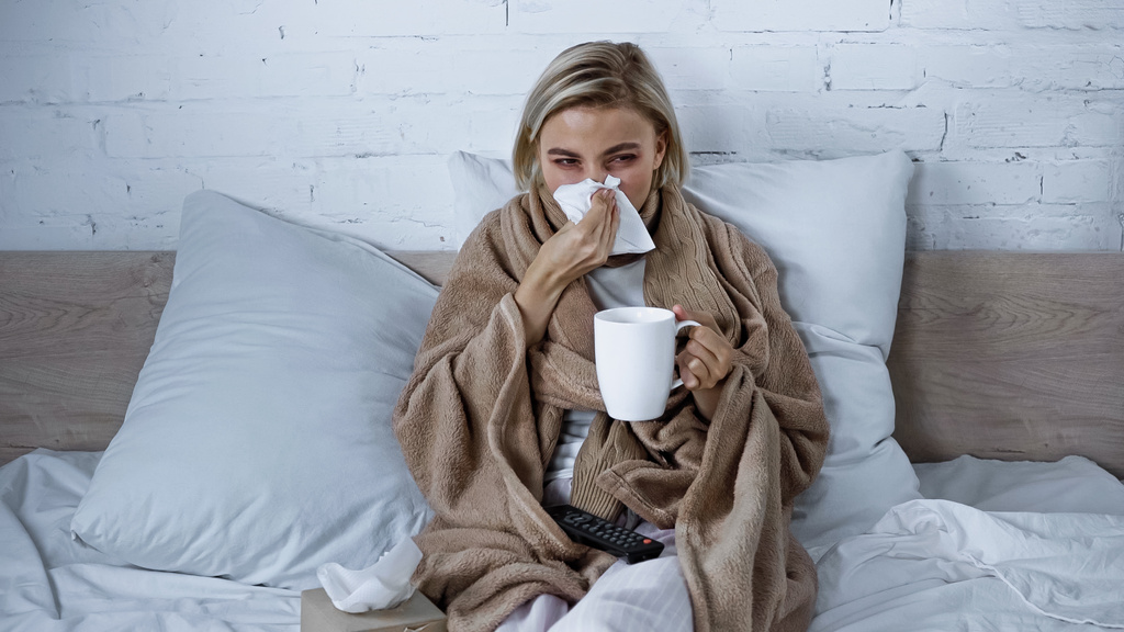 sick woman sneezing into paper napkin while watching tv in bedroom with cup of tea - Photo, Image
