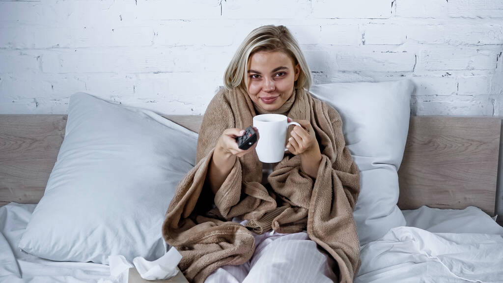 хвора, усміхнена жінка натискає телевізійні канали, тримаючи чашку чаю в спальні
 - Фото, зображення