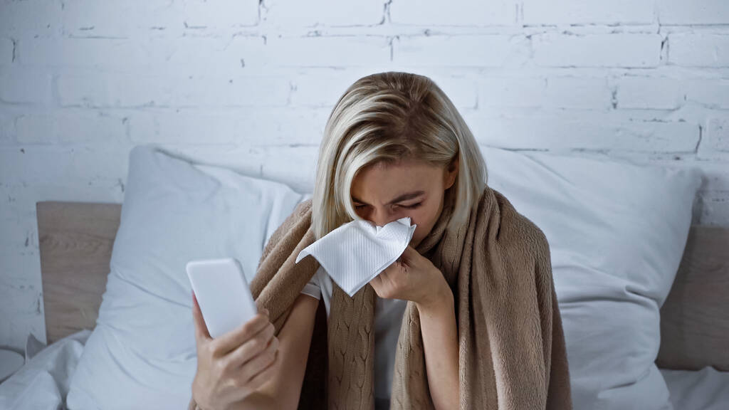 nemocná žena kýchání do papírového ubrousku při držení smartphone v ložnici - Fotografie, Obrázek