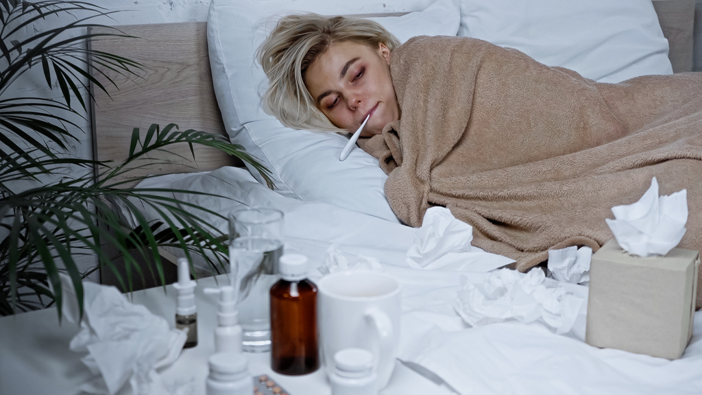 zieke vrouw meten temperatuur in bed in de buurt van servetten en medicijnen op wazig voorgrond - Foto, afbeelding