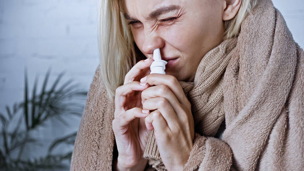 mujer joven enferma frunciendo el ceño mientras usa aerosol nasal  - Foto, Imagen