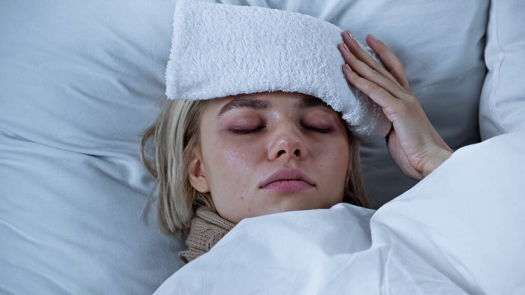 Kranke Frau liegt mit geschlossenen Augen und berührt Kompresse auf der Stirn - Foto, Bild