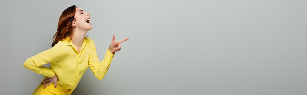 donna eccitata ridendo e indicando con il dito mentre in piedi con mano sul fianco su grigio, banner - Foto, immagini