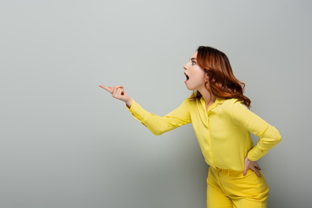 schockierte Frau zeigt mit dem Finger, während sie mit der Hand auf der Hüfte auf grau steht - Foto, Bild
