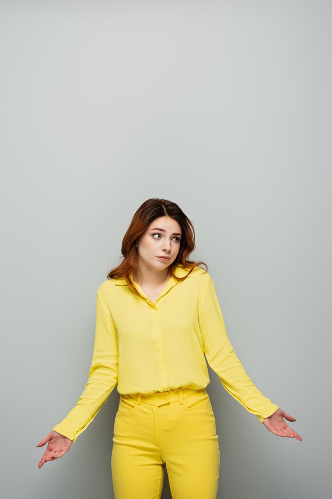 mujer confusa en blusa amarilla mirando hacia otro lado y mostrando gesto de encogimiento en gris - Foto, Imagen