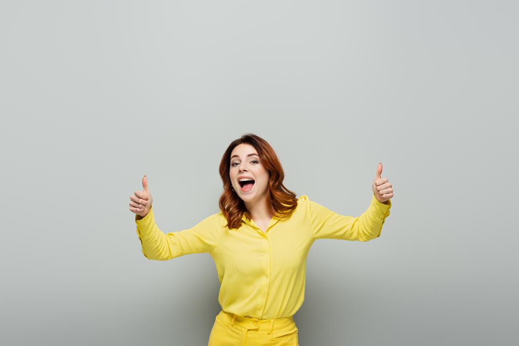 χαρούμενη γυναίκα που δείχνει τους αντίχειρες όρθιες ενώ στέκεται με ανοιχτό το στόμα στο γκρι - Φωτογραφία, εικόνα