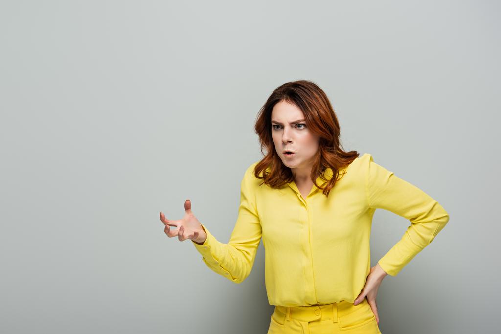 сердитая женщина показывает пугающий жест, стоя с рукой на бедре на сером - Фото, изображение