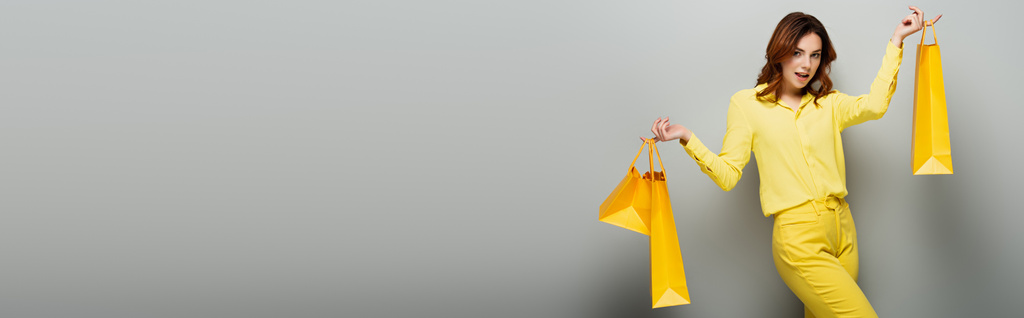 mujer alegre mirando a la cámara mientras sostiene bolsas amarillas en gris, pancarta - Foto, imagen