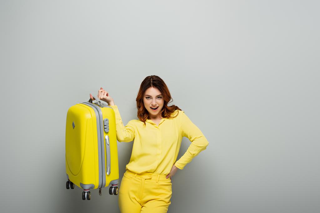 ενθουσιασμένοι γυναίκα κρατώντας κίτρινη βαλίτσα, ενώ στέκεται με το χέρι στο ισχίο σε γκρι - Φωτογραφία, εικόνα