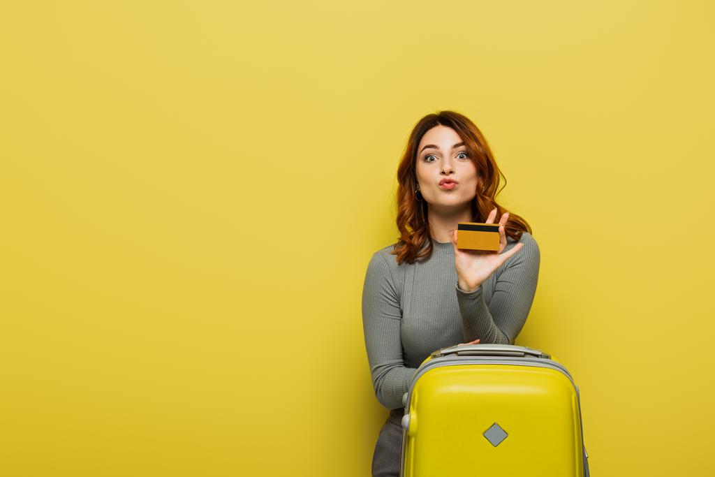 femme aux cheveux bouclés boudant lèvres tout en tenant bagages et carte de crédit sur jaune - Photo, image