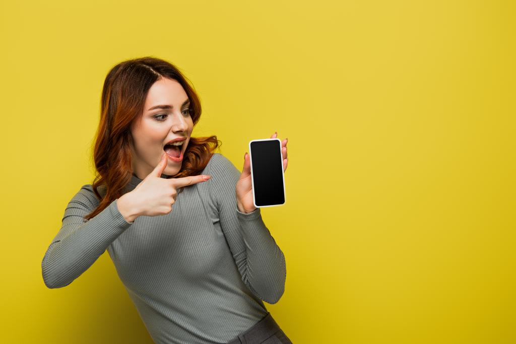 κατάπληκτη γυναίκα με σγουρά μαλλιά που δείχνουν με το δάχτυλο στο smartphone με λευκή οθόνη στο κίτρινο - Φωτογραφία, εικόνα