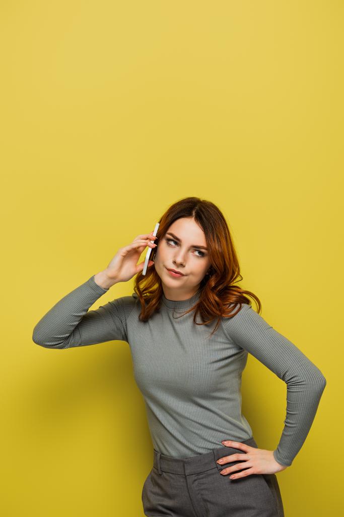 znuděná mladá žena s kudrnatými vlasy mluví na smartphonu a stojí s rukou na boku na žluté - Fotografie, Obrázek