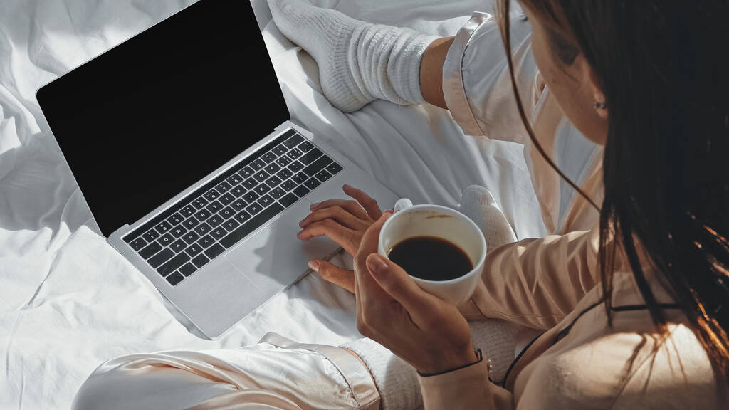vista de ángulo alto de la mujer sosteniendo la taza de café cerca del ordenador portátil con pantalla en blanco en la cama - Foto, Imagen