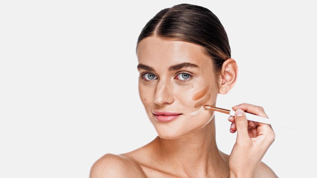 Young adult woman applying different shades of concealer on cheek isolated on white - Photo, Image