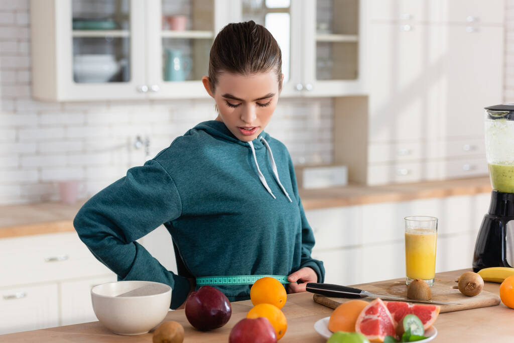 молодая женщина измеряет талию рядом со свежими фруктами на кухонном столе - Фото, изображение