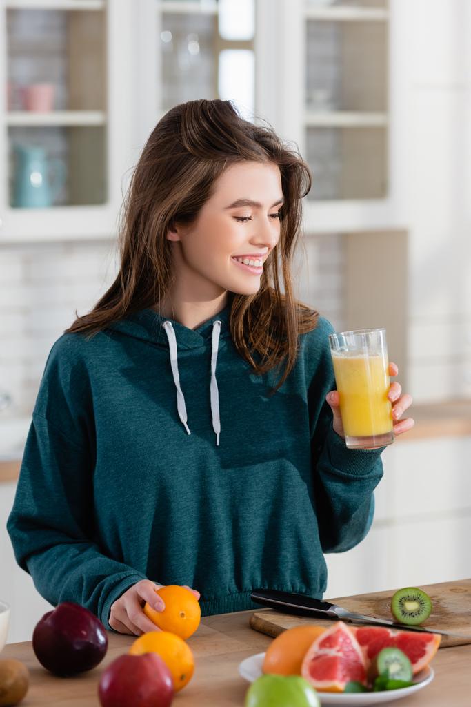 Fröhliche Frau hält frischen Smoothie neben Obst auf Küchentisch - Foto, Bild