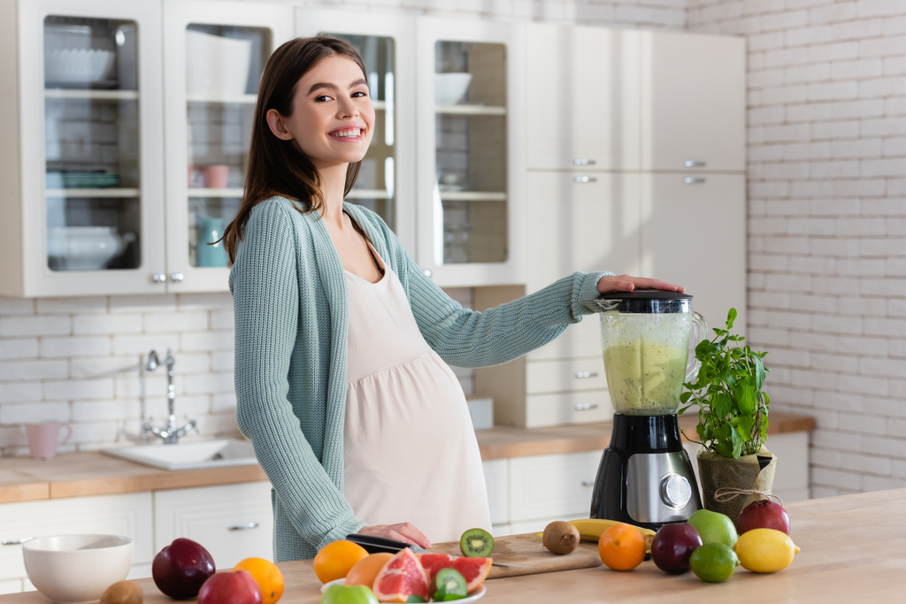 Schwangere lächelt in die Kamera, während sie in der Küche Frucht-Smoothie zubereitet - Foto, Bild