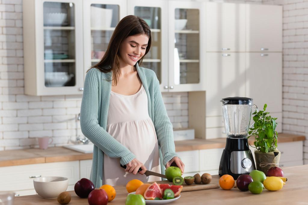 vidám terhes nő vágás friss gyümölcsök közelében turmixgép a konyhában - Fotó, kép