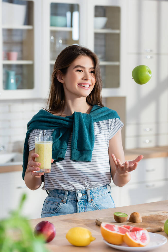 veselá žena žonglovat s jablkem, zatímco drží čerstvé smoothie na rozmazané popředí - Fotografie, Obrázek