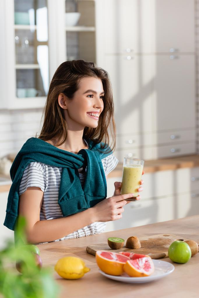 mujer alegre mirando hacia otro lado mientras sostiene el vaso de batido cerca de frutas frescas, borrosa primer plano - Foto, Imagen