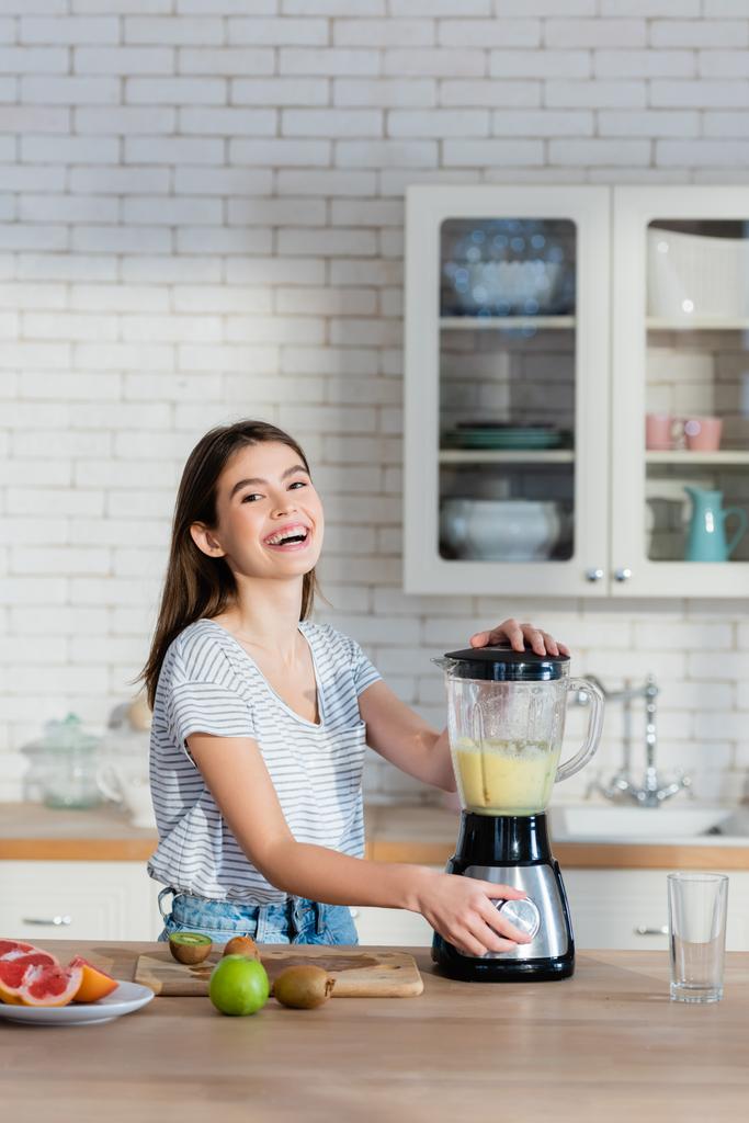 γελώντας γυναίκα κοιτάζοντας κάμερα, ενώ την προετοιμασία smoothie φρούτων στην κουζίνα - Φωτογραφία, εικόνα