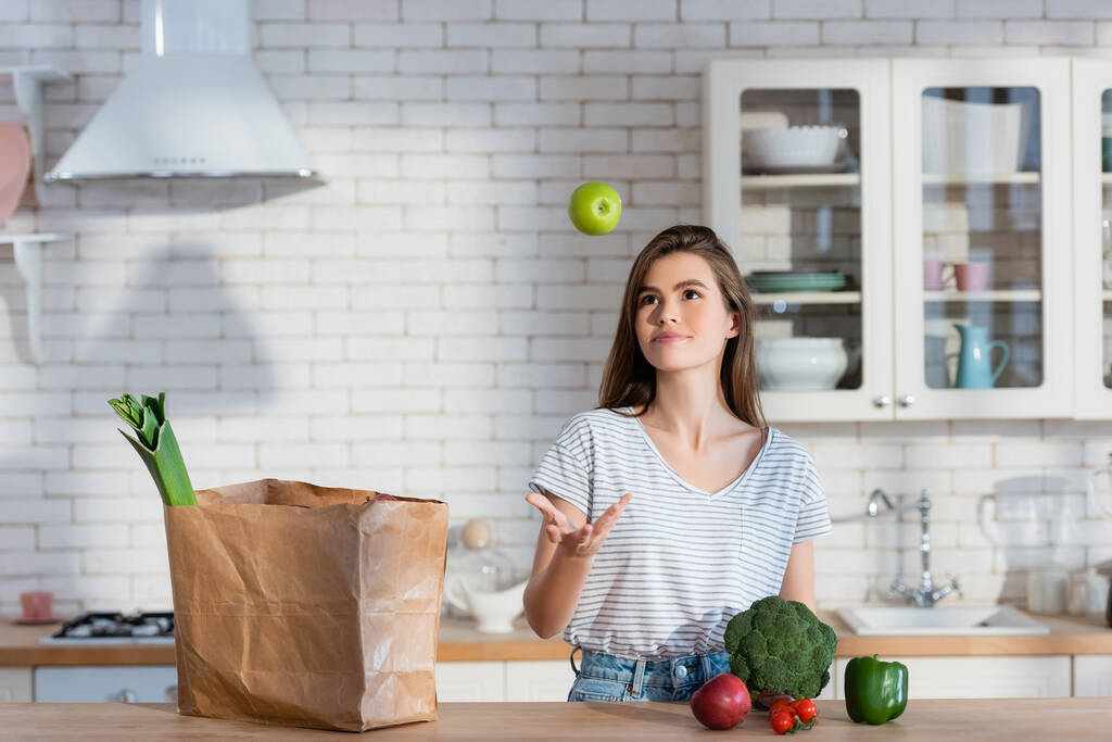 hymyilevä nainen jongleeraa kypsä omena lähellä paperipussi ja tuoreita vihanneksia keittiössä - Valokuva, kuva