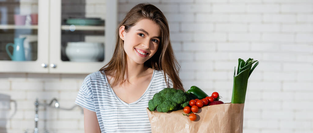 χαρούμενη γυναίκα κοιτάζοντας κάμερα κοντά σε φρέσκα λαχανικά σε χάρτινη σακούλα, πανό - Φωτογραφία, εικόνα