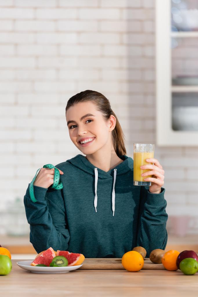 весела жінка тримає мірну стрічку і склянку фруктового соку на кухні
 - Фото, зображення