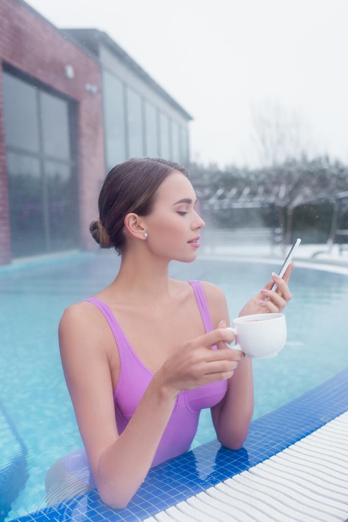 mujer joven sosteniendo teléfono inteligente y taza mientras se baña en la piscina de aguas termales - Foto, imagen