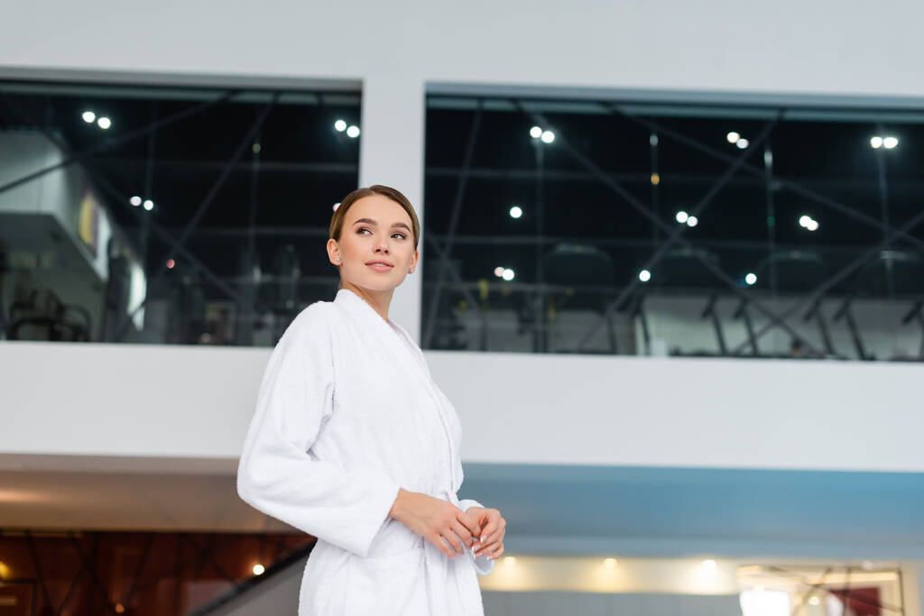 mujer joven sonriente en albornoz blanco de pie en el centro de spa  - Foto, Imagen
