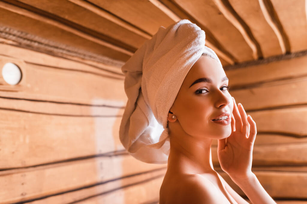 Glimlachende vrouw met handdoek op hoofd kijkend naar camera in sauna op wazige achtergrond  - Foto, afbeelding