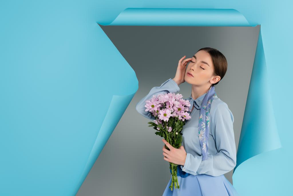 elegante Frau, die das Gesicht berührt, während sie mit geschlossenen Augen und rosa Blumen neben blauem zerrissenem Papier auf grauem Hintergrund posiert - Foto, Bild
