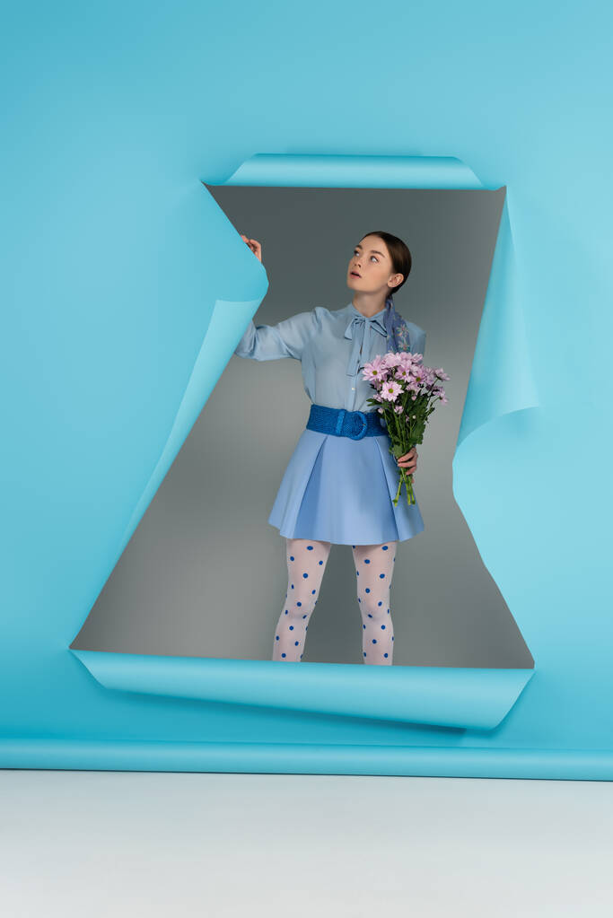 fiatal divatos nő pontozott harisnyanadrág áll a virágok közelében lyuk kék papír szürke háttér - Fotó, kép