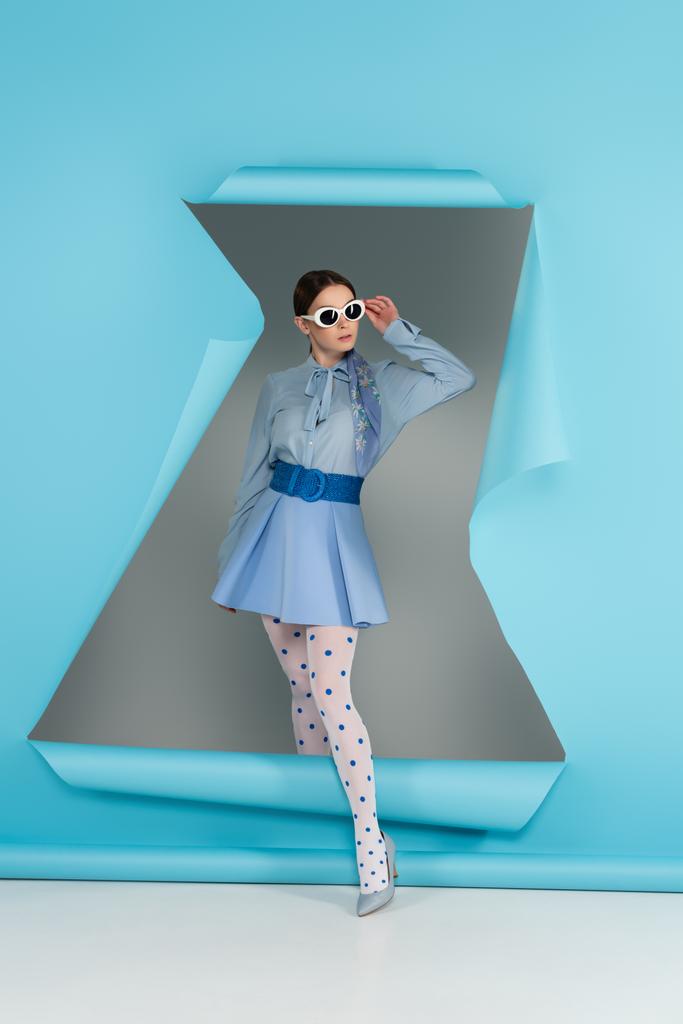 giovane donna in abiti glamour regolazione occhiali da sole vicino foro in carta blu su sfondo grigio - Foto, immagini